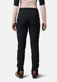 Pantalons de ski noirs avec une coupe ajustée, des poignets élastiques et deux poches arrière. Portés avec des bottes de ski noires et orange.