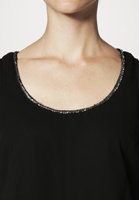 Haut noir avec un col rond orné d'une bordure en sequins argentés. Le tissu semble lisse et léger.