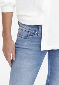 Jeans de mezclilla azul claro con un ajuste slim, que presentan un diseño clásico de cinco bolsillos y detalles de costura sutiles. Complementados con una camisa blanca texturizada.