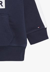 Sudadera azul marino con detalles blancos, puños y dobladillo acanalados, bolsillo tipo canguro y logo en la parte delantera. Textura suave y diseño informal.