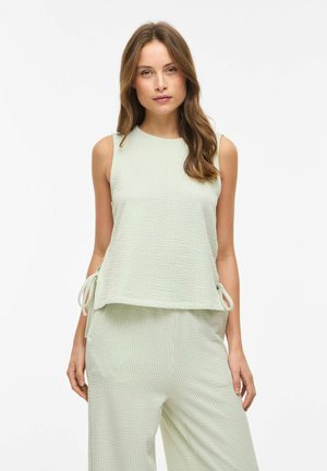 Kvinde iført ærmeløs, lysegrøn struktureret top og matchende bukser med bindedetaljer i siden, stående mod en ensfarvet hvid baggrund.