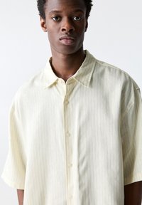 Chemise à manches courtes, de couleur jaune clair, avec des rayures verticales, une coupe décontractée et un col soigné ; le tissu semble lisse et légèrement texturé.