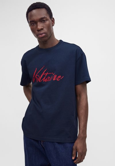 Νεαρός άνδρας που φοράει μπλε σκούρο t-shirt με κόκκινο κείμενο "Voltaire" και σκούρο μπλε τζιν, στέκεται μπροστά σε ένα απλό φωτεινό φόντο.