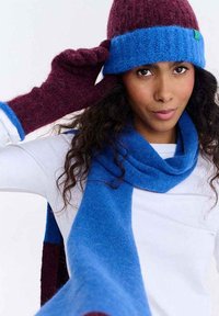 Kapelusz, szalik i rękawiczki wykonane z miękkiej wełny. Kapelusz ma bordowy korpus z niebieskim paskiem. Szalik i rękawiczki są niebieskie z bordowymi akcentami.