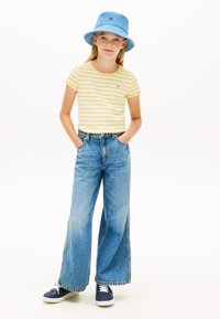 Ριγέ κίτρινο και λευκό τοπ με κοντά μανίκια και κυματιστά τελειώματα, σε συνδυασμό με μπλε τζιν παντελόνι φαρδιάς γραμμής και μπλε καπέλο bucket. Αθλητικά παπούτσια με λευκά κορδόνια.