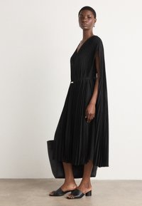 Black pleated dress with a V-neck, cape sleeves, and a waist belt. Paired with black open-toe mules and a large black tote bag.