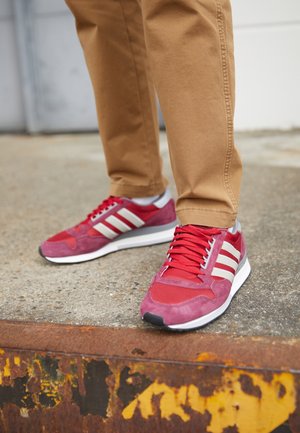 Personne portant un pantalon beige et des baskets rouges avec des rayures blanches, debout sur une surface en béton près d'un bord jaune rouillé.