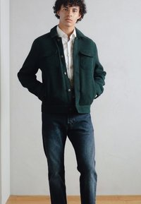 Giacca verde con texture, dotata di tasche frontali, chiusura a bottoni e colletto, indossata sopra una camicia bianca, abbinata a jeans scuri. Sfondo neutro.