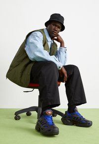 Svart och blå sneakers med utskärningar, ihop med svarta byxor, en denimskjorta och en olivgrön pufferväst. Svart bucket hat. Grön golv.