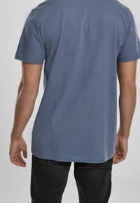 Muž oblečený v jednoduchém modrém tričku s krátkým rukávem a černých kalhotách, zobrazený zezadu na neutrálním pozadí.