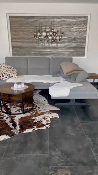 Divano sezione grigia con cuscini, coperta bianca e un tavolino da caffè rotondo in legno. Tappeto in pelle di mucca texture su pavimento piastrellato scuro. Arte murale sopra.