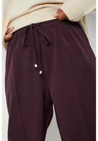 Dunkelviolette, leichte, weiche Stoffhosen mit einem elastischen Bund und Kordelzug. Silbernene Metallenden betonen die Kordeln.