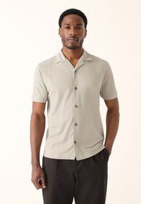 Kortärmad, ljust grå knappskjorta med campkrage, slät textur och mörka knappar, bärs ledigt över mörka byxor.