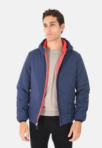 Giacca a vento navy con una fodera interna rossa, cappuccio, chiusura frontale con zip e polsini elasticizzati. Materiale texturizzato e isolato per il calore.
