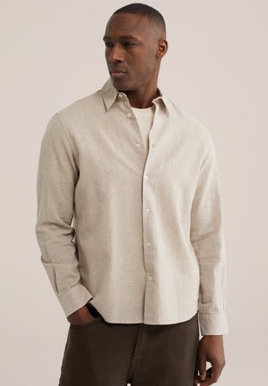 Muž nosiaci svetlobéžovú textúrovanú košeľu na gombíky cez biele tričko, s tmavými nohavicami, stojaci s jednou rukou v vrecku, pozerajúci sa bokom.