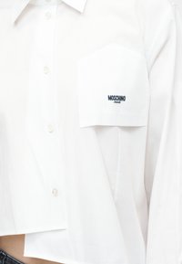Λευκό πουκάμισο με κουμπιά και τσέπη στο στήθος, κεντημένη με τη φράση «MOSCHINO JEANS» με μαύρο νήμα.