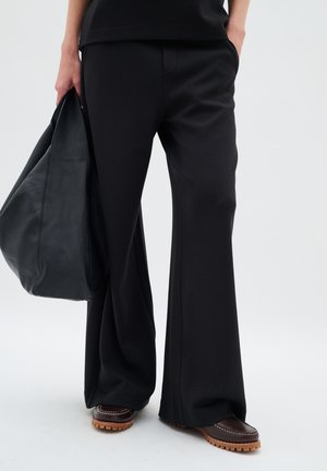 Black, flared trousers made of soft fabric, featuring a front pocket and side seams. Paired with brown loafers and a black leather tote bag.