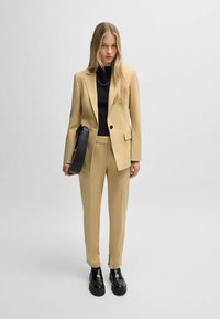 Beige tailored suit, black top, and black shoulder bag. Smooth texture, structured fit, and a single button on the blazer. Chunky black shoes.