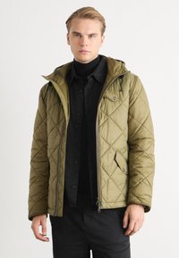 Veste matelassée vert olive avec fermeture éclair à l'avant, capuche et poches à pression. Texture lisse, motif en diamant, portée sur une chemise noire.
