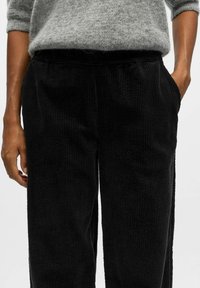 Pantaloni di velluto a coste neri con texture a righe verticali, dotati di tasche laterali e una cintura elastica. Design minimalista senza decorazioni.