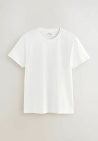 Simpel hvid t-shirt med korte ærmer og rund halsudskæring, med en medium størrelse etiket, hængende mod en lys grå væg.