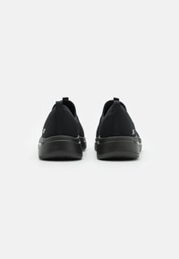 Zapatillas deportivas negras con parte superior de malla, diseño elástico y suela acolchada. Presenta una pestaña para tirar en el talón y un sutil acento de logo.