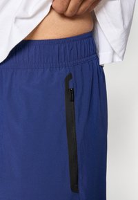 Calções atléticos azuis com cintura elástica, apresentando um bolso lateral com fecho preto e tecido de textura suave.