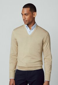 Beige V-hals trui van zachte stof. Kenmerkt lange mouwen, geribbelde manchetten en een gladde textuur. Draagt over een lichtblauw overhemd met kraag.