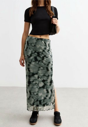 Femme portant un t-shirt noir court, une jupe longue tie-dye verte et noire avec une fente sur le côté, des baskets noires, et tenant un petit sac à épaule noir.