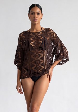 Femme portant un haut en crochet marron sur un bas de bikini noir, debout devant un fond clair uni.