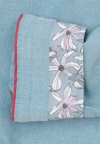 Lichtblauwe stof met een gerolde rand, versierd met een bloemenprint strook en een dunne rode piping langs de rand.