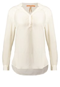 Bílá dlouhá slavnostní blůza s výstřihem do V, částečnou přední knoflíkovou léga, zakřiveným spodním lemem a lehkým texturovaným materiálem.