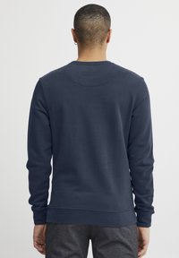 Tamsiai mėlyna džemperis su apvaliu kaklu, ilgomis rankovėmis ir banguotu apdailu. Lygi tekstūra, su minimaliais siuvimo akcentais.