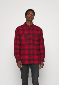 Red and black plaid flannel shirt with long sleeves, button closure, and two chest pockets. Features a pointed collar and textured fabric.