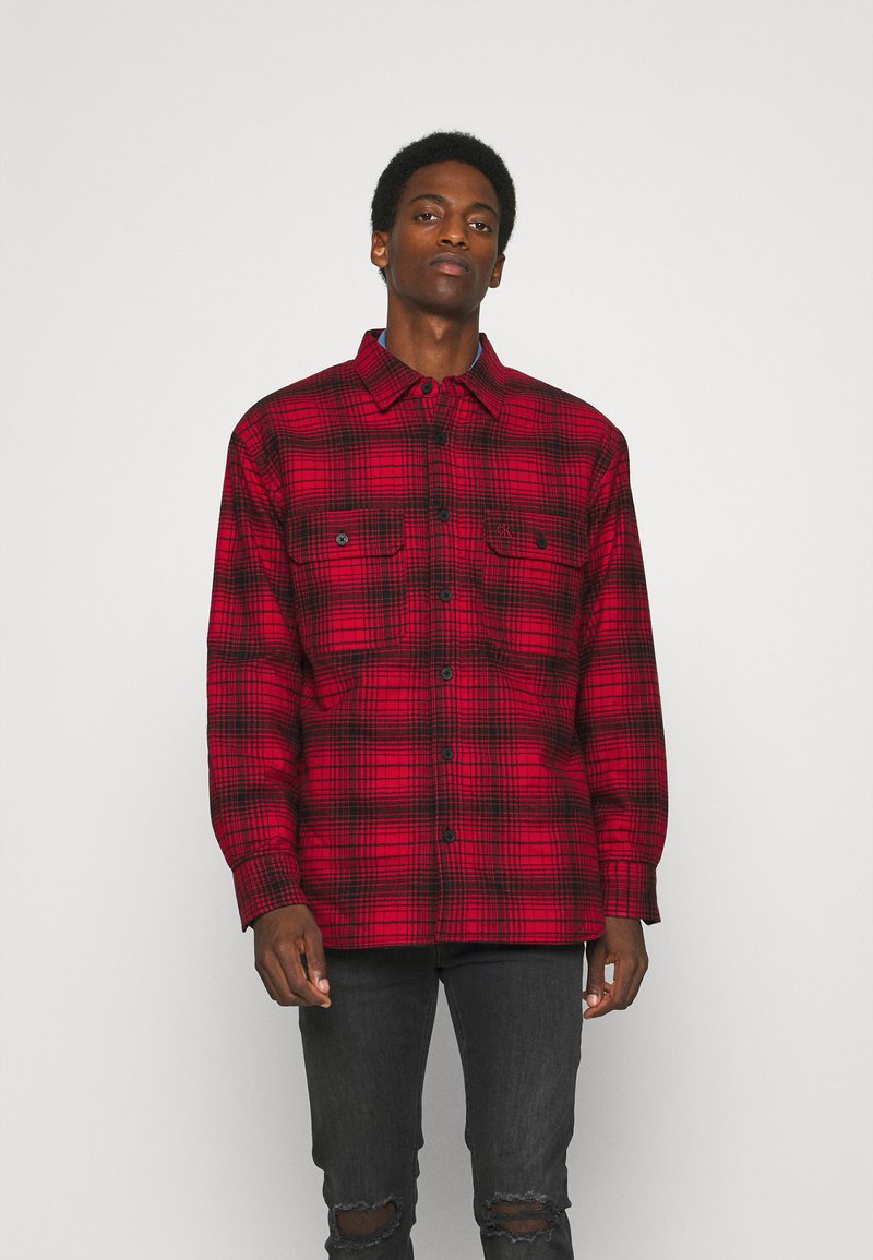Red and black plaid flannel shirt with long sleeves, button closure, and two chest pockets. Features a pointed collar and textured fabric.