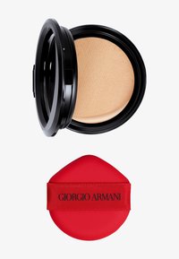 Fond de teint compact dans un étui noir avec une surface beige texturée. Comprend une éponge applicatrice rouge avec une sangle de logo noire.