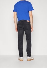 Mörka slim-fit jeans med bakfickor, parat med en blå bomulls-t-shirt. Vita och beige sneakers, synlig sömnad och detalj med varumärkeslogotyp.