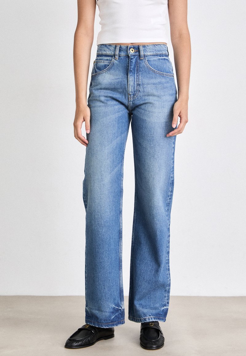 Jeans de denim azul claro con cintura alta, piernas anchas y bolsillos frontales. Presentan una cremallera y trabillas para cinturón, con un desgaste mínimo.