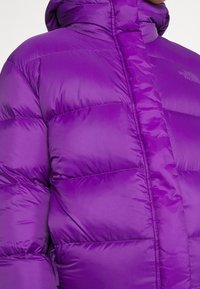 Violetti puhallettava takki korkealla kauluksella, tikattu muotoilu, sileä pinta ja logoyksityiskohta vasemmalla ylhäällä.