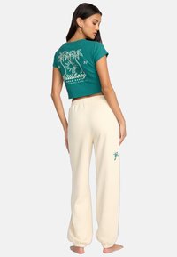 Zielony krótki t-shirt z graficznym motywem palmy i białym napisem, zestawiony z kremowymi spodniami dresowymi z małym wzorem palmy.