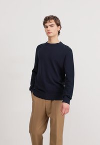 Maglione blu navy a maglia con scollatura rotonda e polsini a costine, abbinato a pantaloni beige chiaro su misura. Tessuto morbido, vestibilità classica.