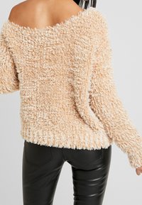 Maglione beige peloso con design off-shoulder, maniche lunghe e una consistenza morbida. Indossato con pantaloni neri aderenti.