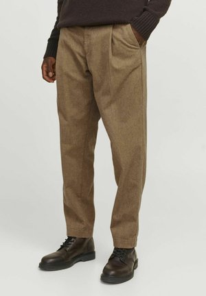 Mężczyzna w brązowych plisowanych spodniach, czarnych sznurowanych butach i ciemnym swetrze, stojący z jedną ręką w kieszeni na jednolitym tle.