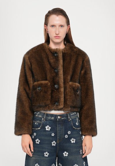 Persona llevando una chaqueta de pelaje sintético marrón oscuro corta con grandes botones negros y unos pantalones vaqueros de denim azul oscuro con estampado floral.