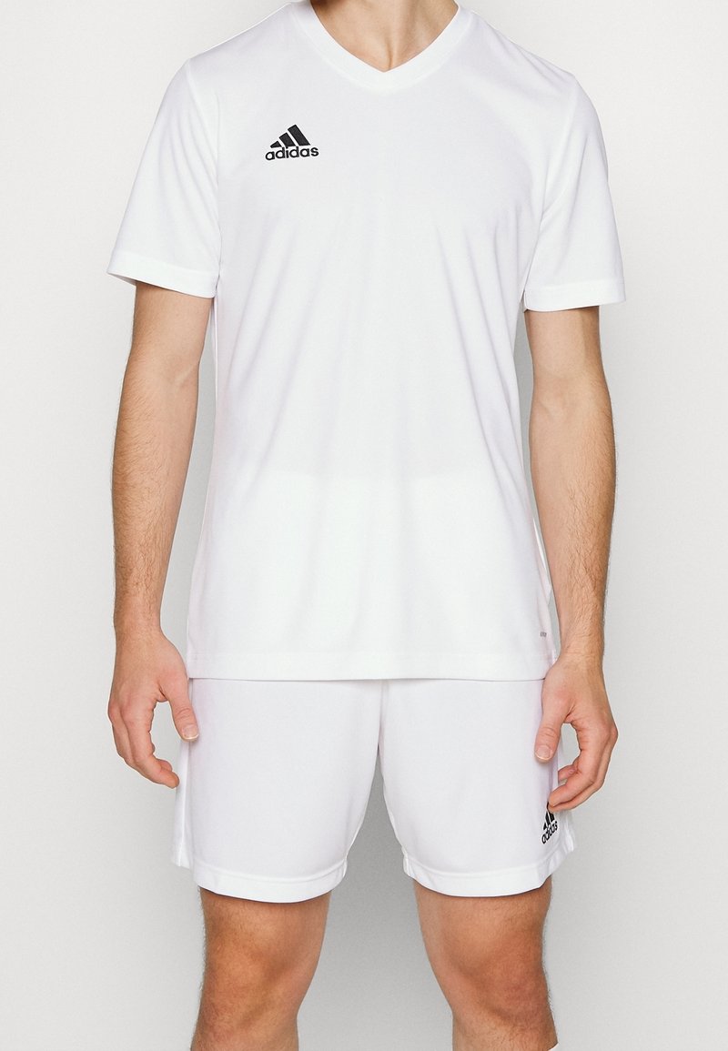 Man som bär vit Adidas sportkläder med svart logotyp, kortärmad V-ringad tröja och matchande shorts mot en plain bakgrund.