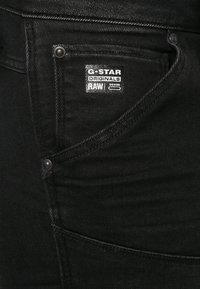 Jeans de mezclilla negra con un primer plano de un bolsillo lateral. Presenta una etiqueta de G-Star Originals y acentos de hardware metálico.