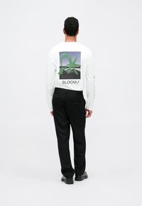 Fehér, hosszú ujjú póló zöld grafikus mintával és "BLOOM/" felirattal a hátsó részén, fekete nadrággal és fekete cipővel párosítva.