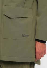 Olijfgroene jas met een gladde textuur, voorzien van een grote voorkant zak en zwarte branding. De manchetten zijn zwart en het ontwerp is strakker en hedendaags.