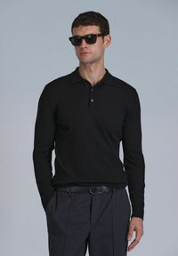 Polo à manches longues côtelé noir avec un col à boutons, associé à un pantalon gris foncé. Le modèle porte des lunettes de soleil noires.