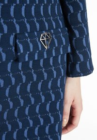 Navy Stoff mit einem strukturierten Muster, das abwechselnd blaue Formen zeigt. Ein silberner diamantförmiger Anhänger akzentuiert eine Tasche des Kleidungsstücks.